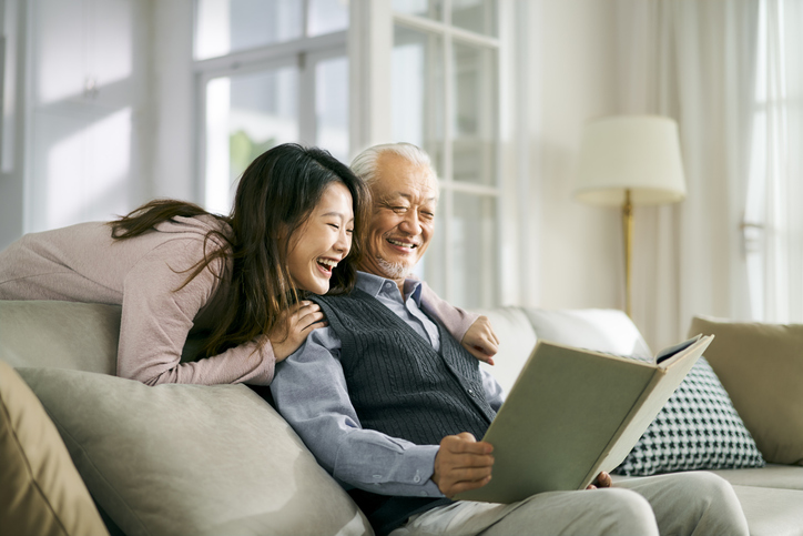 senior man flipping through a book on a couch with his daughter hanging over his shoulders