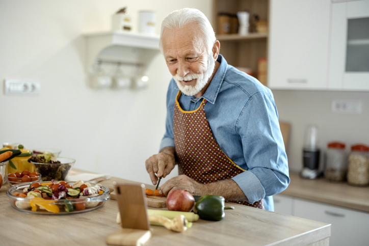 senior man cutting up carrots at his kitchen table while following a recipe on his smartphone